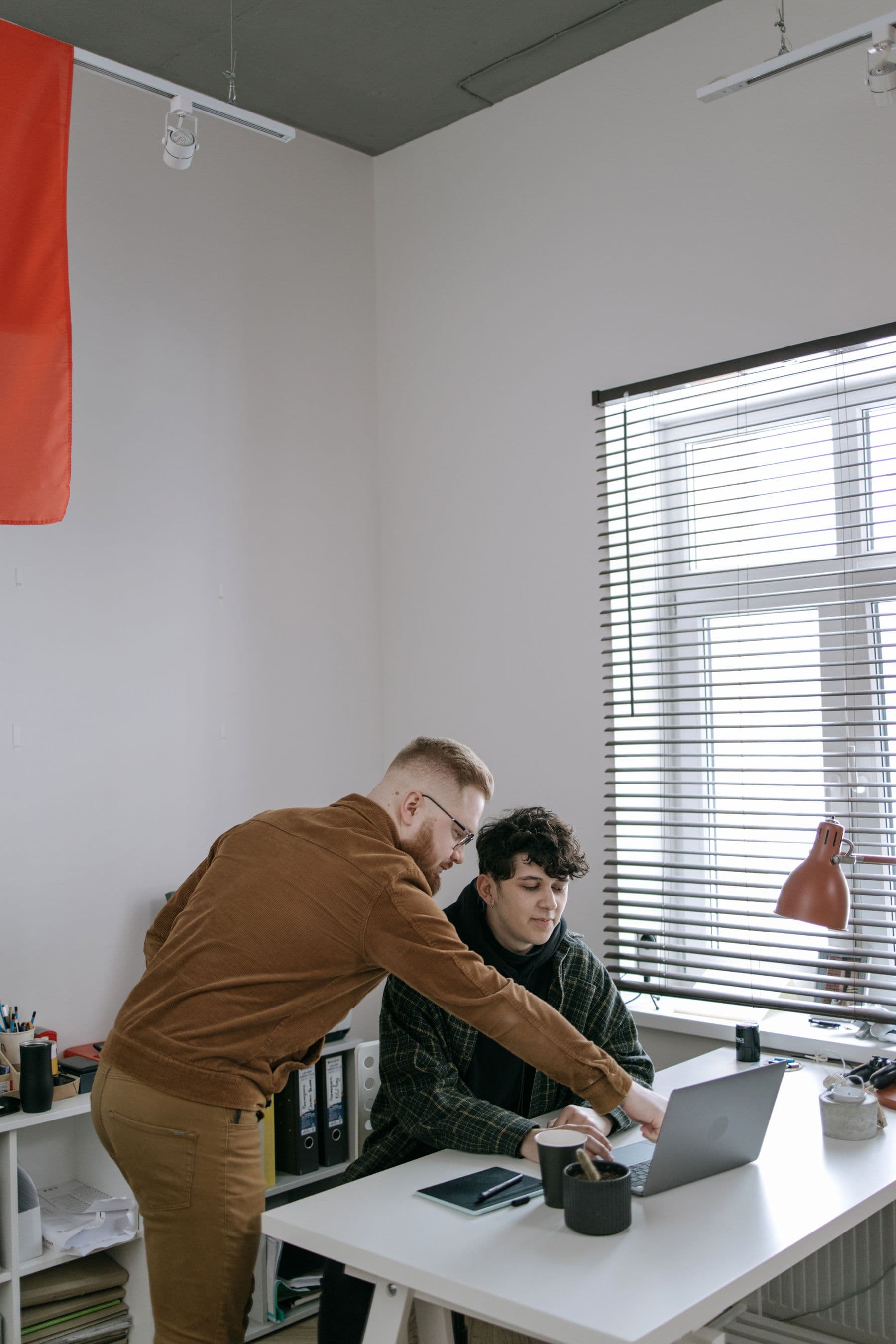 Two men working through a laptop screen at a desk beside a window.
