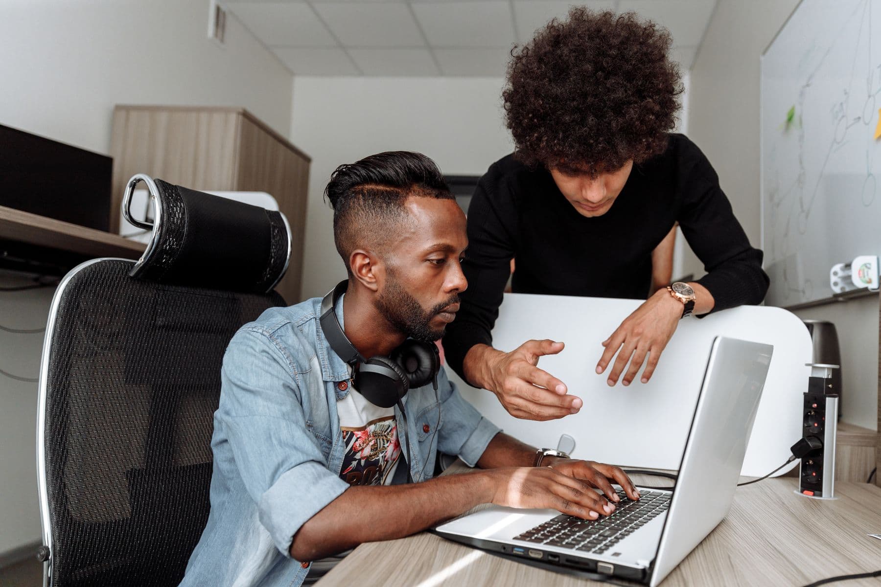 Two men discussing work at a laptop beside a whiteboard.